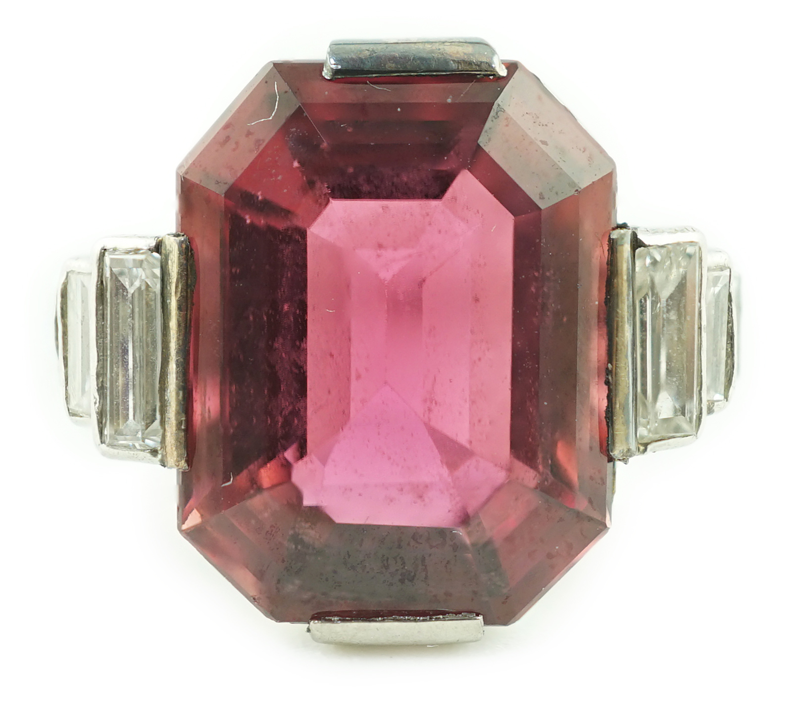 An Art Deco platinum and single stone emerald cut deep pink tourmaline set dress ring, with graduated six stone baguette cut diamond set shoulders